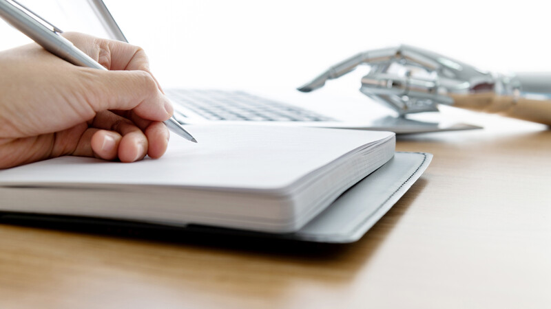 En menneske-hånd skriver med pen mot en notatblokk. En robotarm i bakgrunnen skriver på en PC.