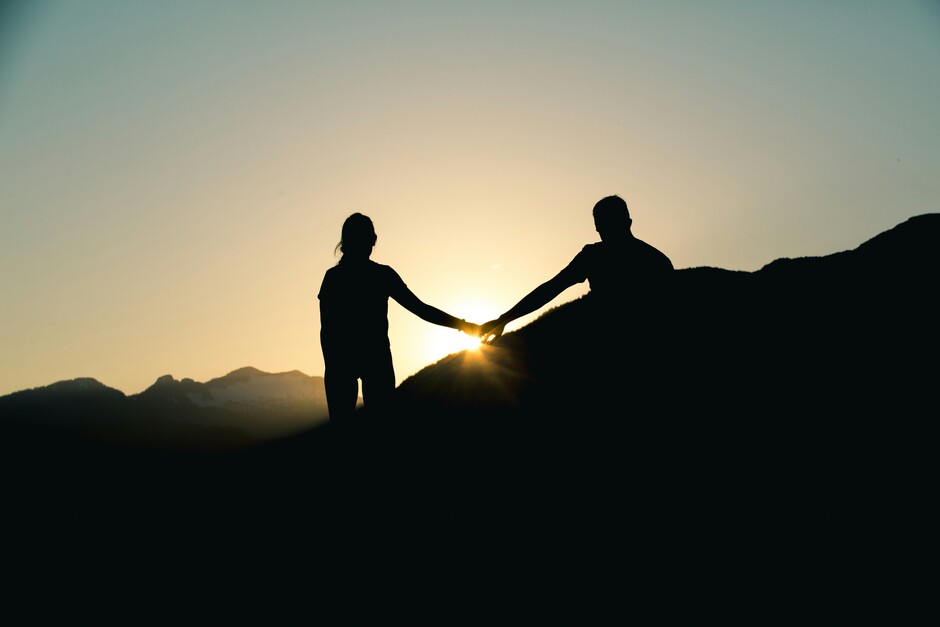 Siluett av to som holder hender i solnedgang på fjelltopp