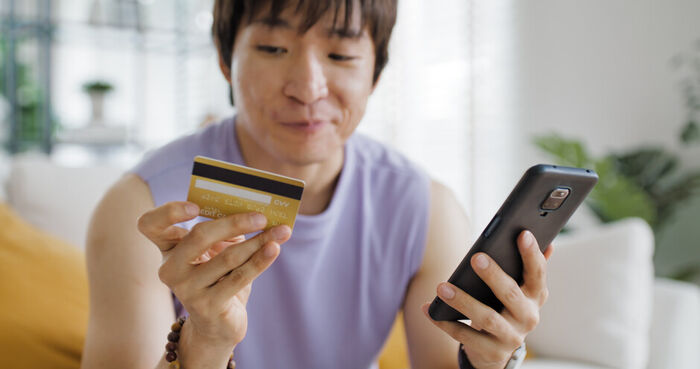 Mann sitter hjemme i stuen med bankkort og mobiltelefon i hendene. Han smiler lurt til bankkortet. 