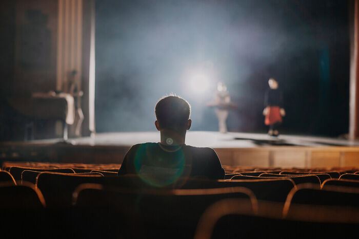 Photo of a person sitting in an auditorium, with someone performing on a stage 