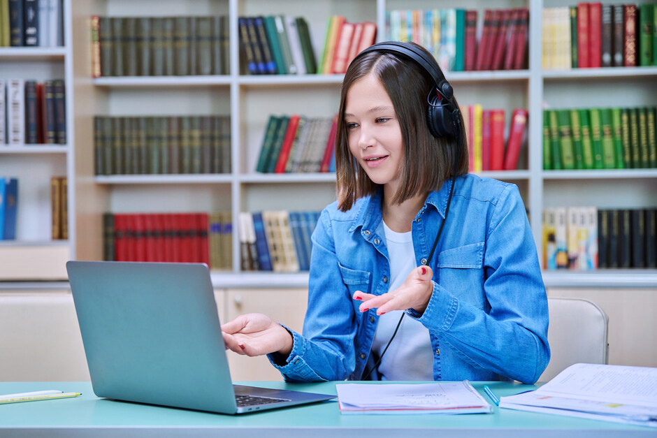 Student sitting in a library, wearing a headset and gesturing at her computer.