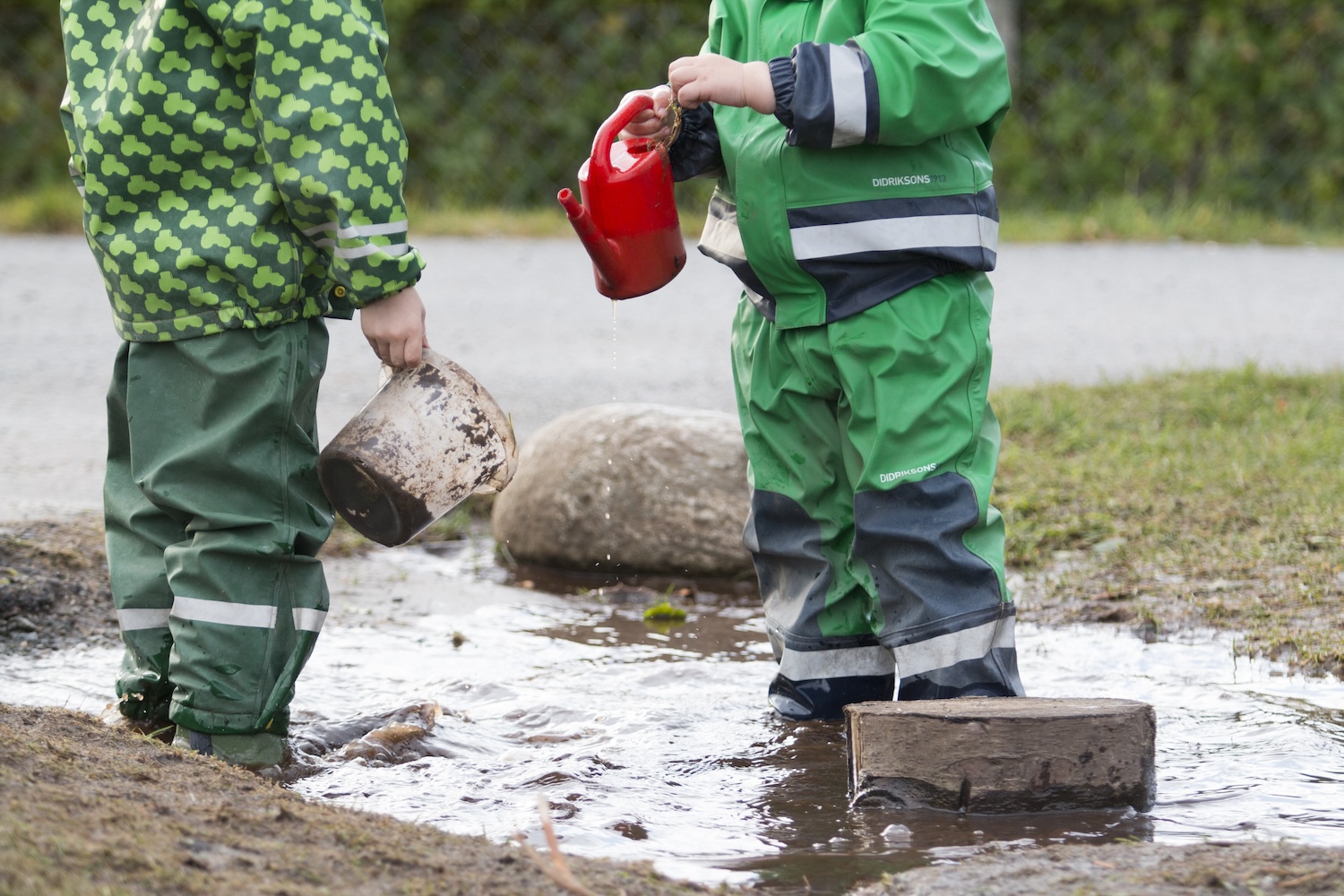 To barn står i sølepytt med regntøy, leker med vannkanne og bøtte