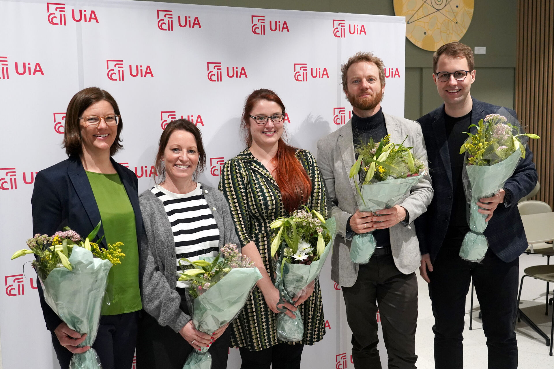 Fem jurymedlemmer står med blomster i hendene foran en plakat med UiA-logoer. 