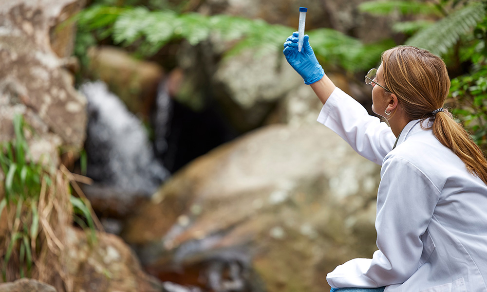Kvinnelig forsker i naturen tester og undersøker vann i en bekk i undersøkelsesglass