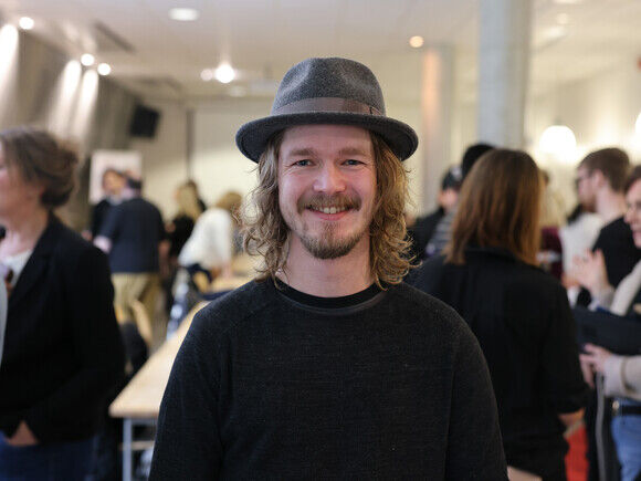 Portrett av studenten Bjørn Kjetil Horverak, ikledd en svart genser og grå hatt. Bjørn har halvlangt hår og smiler til kameraet. 
