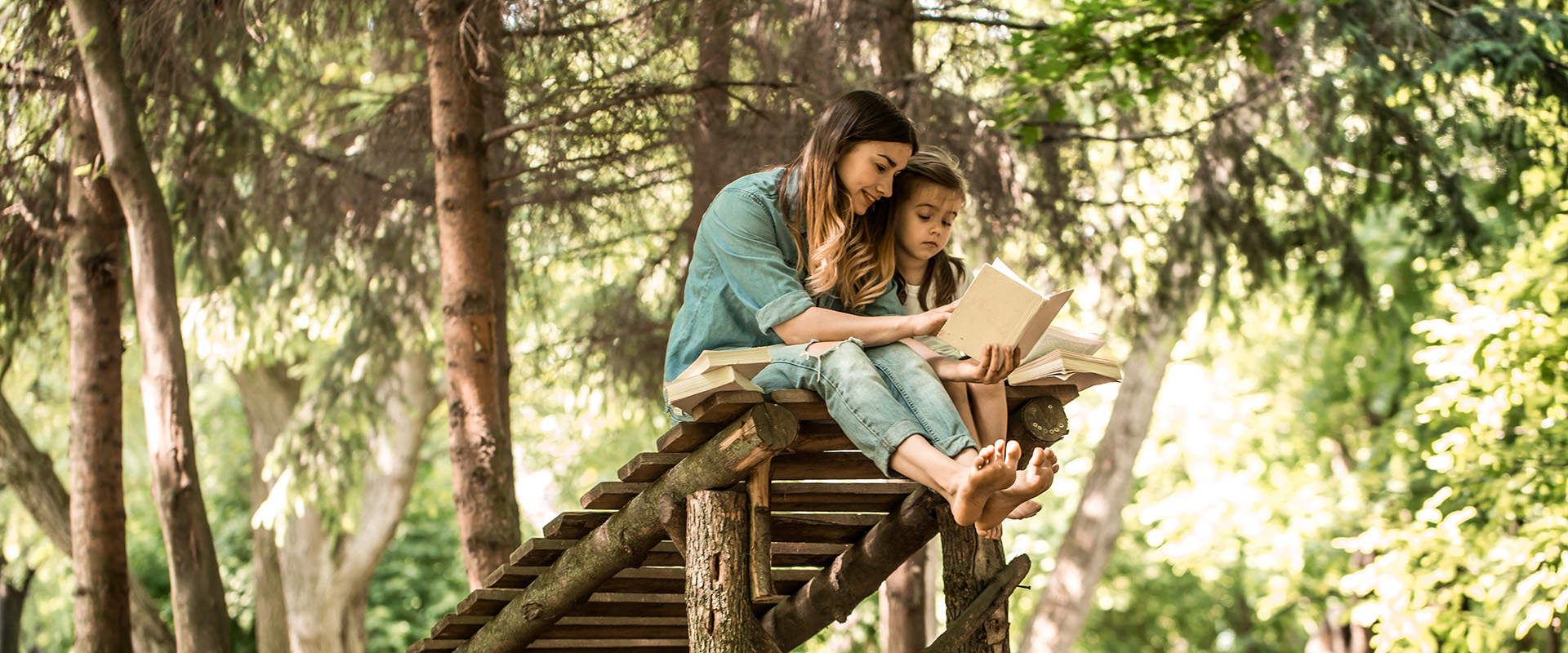 Kvinne og jente sitter på en slags brygge i skogen, leser bok, trær, solglimt gjennom trærne