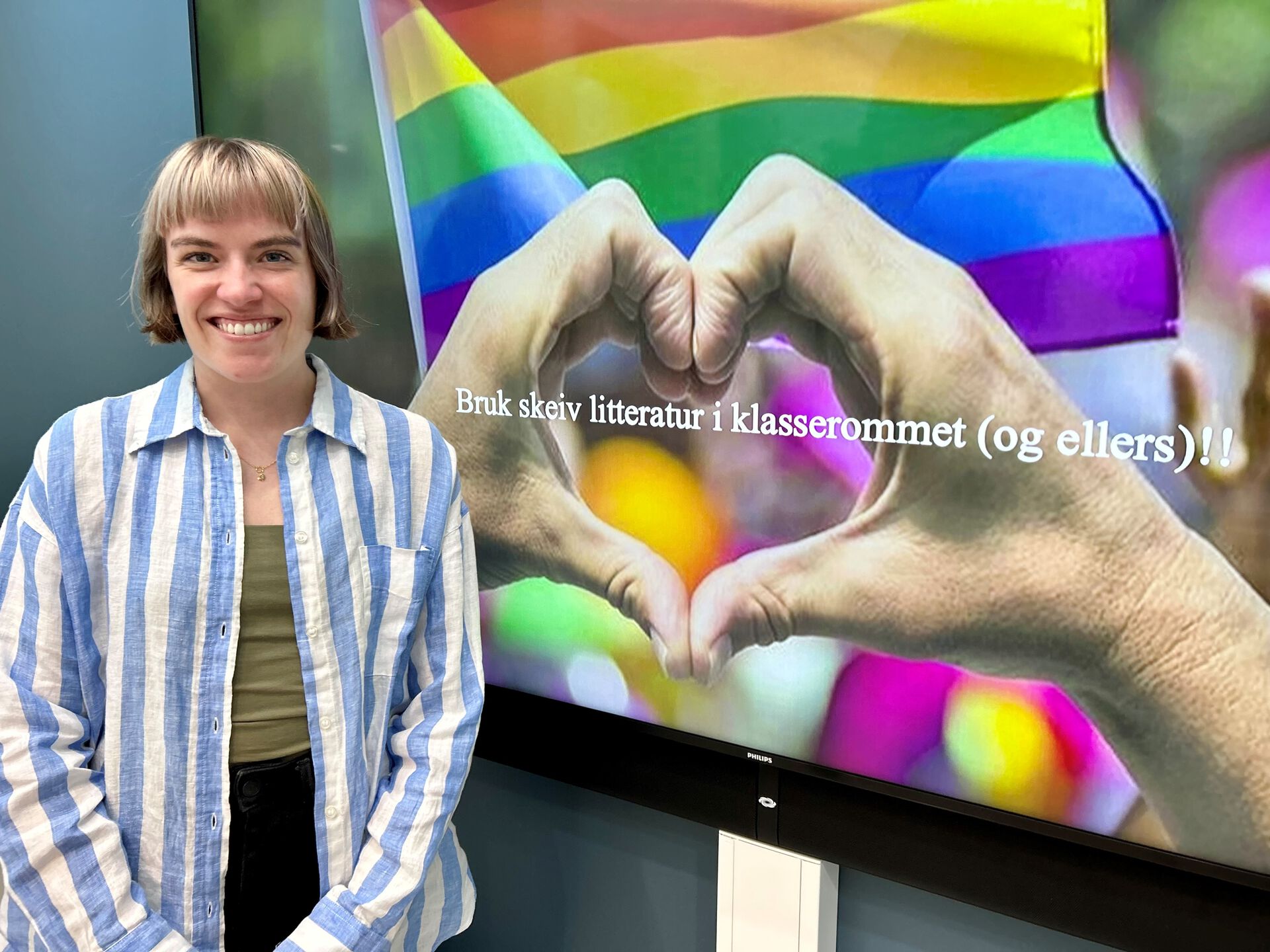 Henrikke Nygaard står foran en stor skjerm. På skjermen er det bilde av to hender som former et hjerte. I bakgrunnen vaier regnbueflagget.
