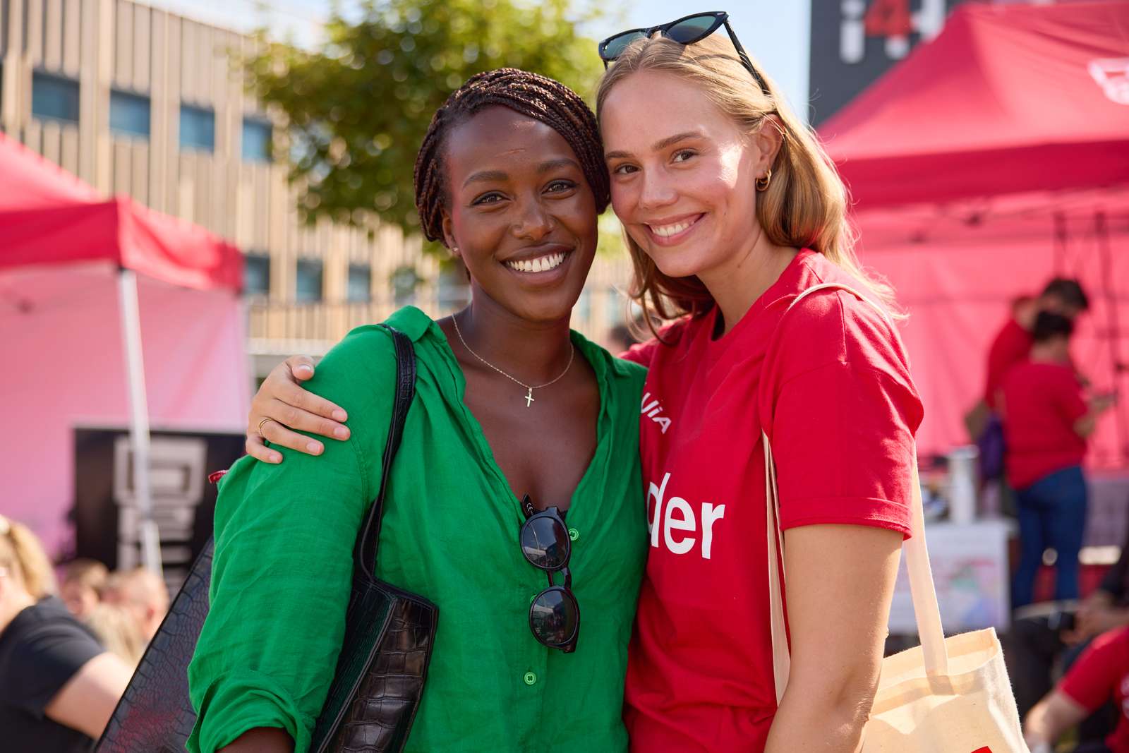 Two women are holding around each other and smiling to the camera. The girl on the left is dark skinned, wearing a green blouse. The girl on the right is light skinned and is wearing a red "buddy" t-shirt.