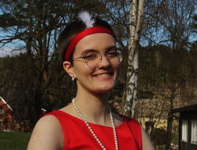Victoria Abelsen poserer smilende i vårsola med koruniformen sin på. Det er en rød kjole med perlesmykke, og rødt hårbånd med en hvit fjær i på hodet.