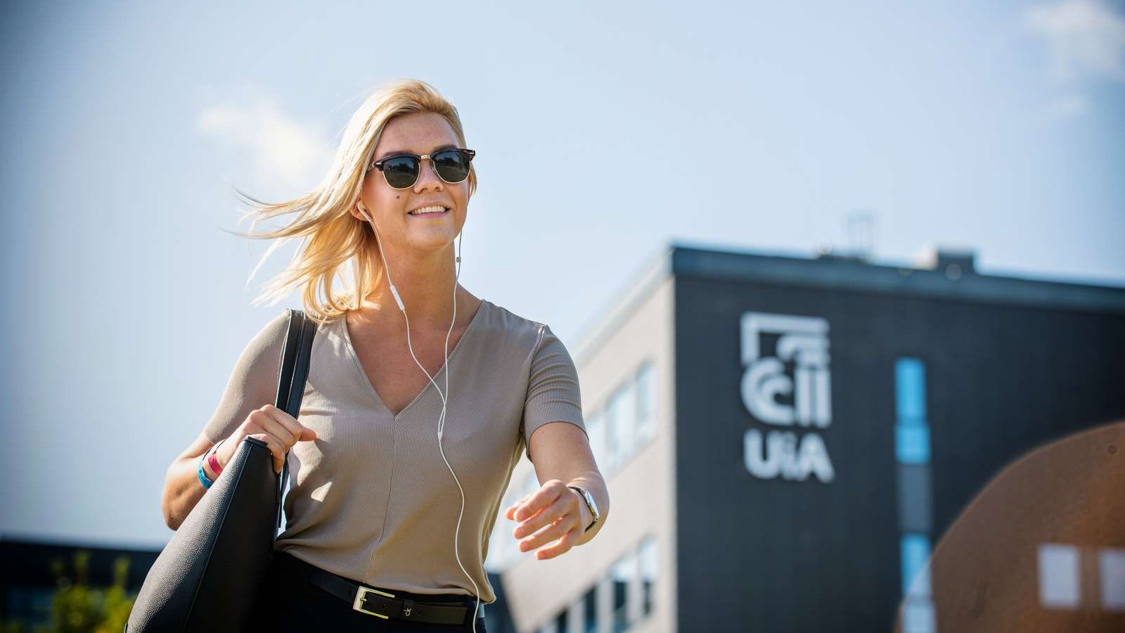 Kvinnelig UiA-student med solbriller, veske og ørepropper som går målrettet med UiAs campus i Grimstad i bakgrunnen. 