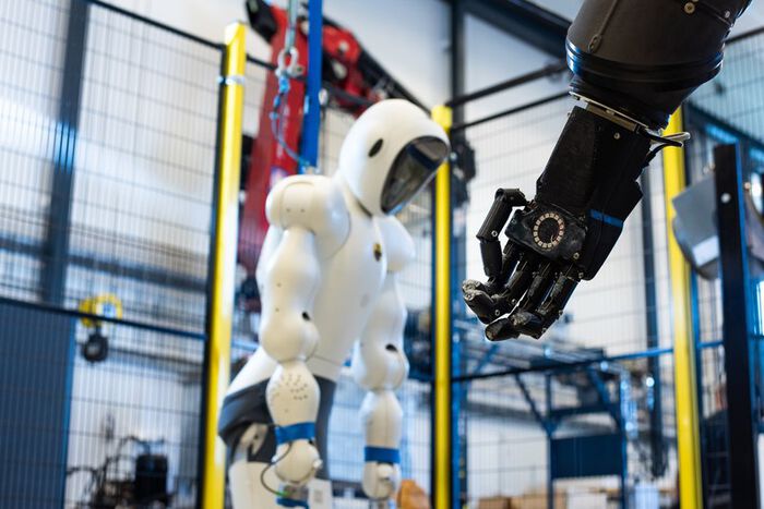 Bildet viser en stor hvit robot som står utstilt på en lab og en svart robothånd i forgrunnen.