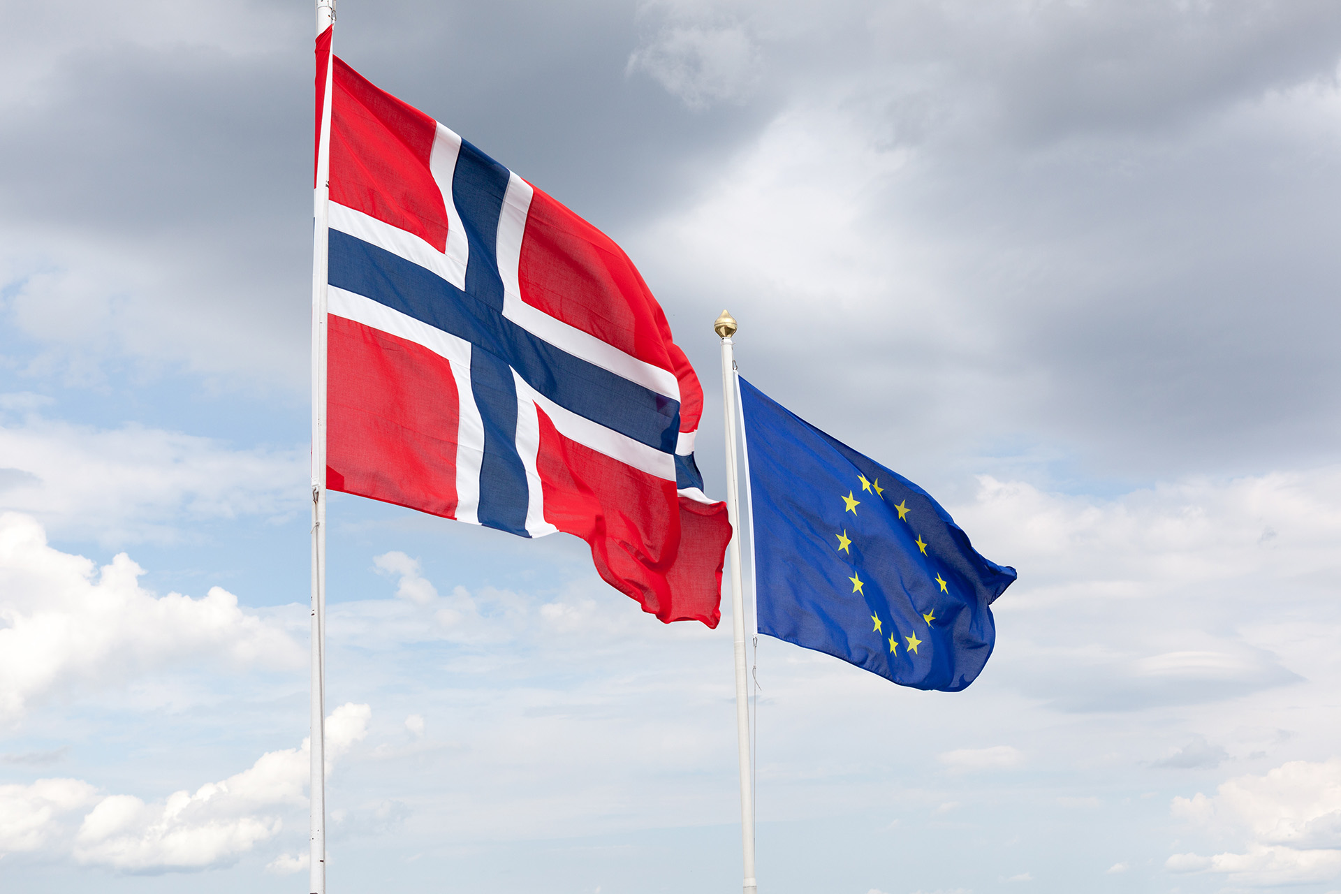 A Norwegian flag waves in the wind with a flag of the European Union in the background against a sky filled with clouds.