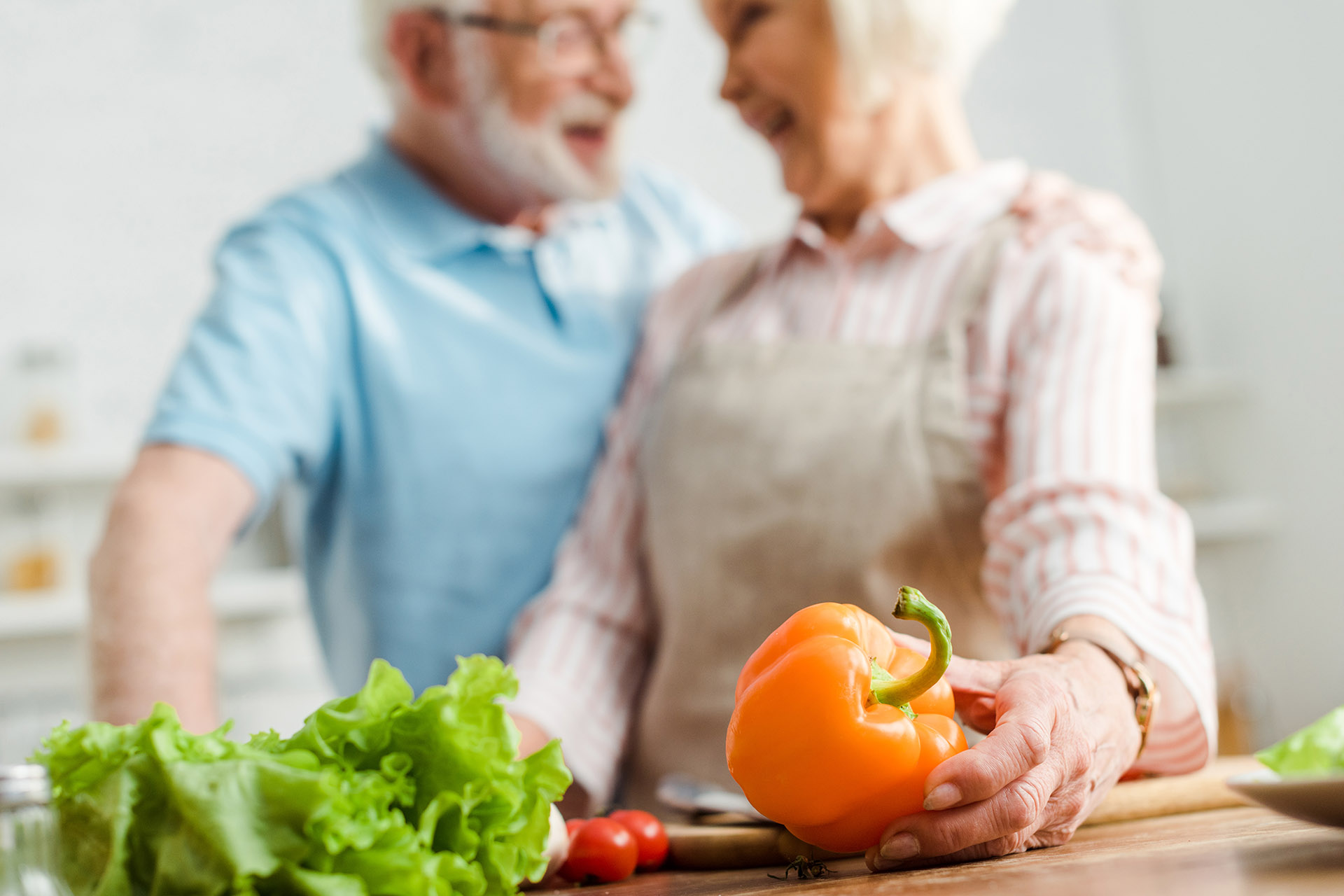 En eldre mann og en eldre kvinne ser på hverandre og smiler bak et bord med grønnsaker 