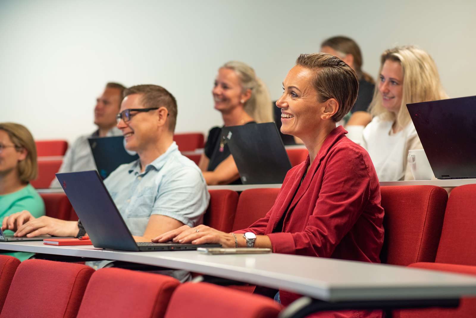 Flere smilende studenter sitter i en forelesningssal med datamaskiner foran seg.
