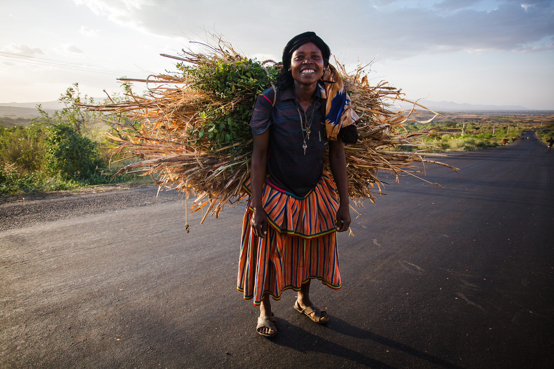 Afrikansk smilende kvinne bærer en bunt med kvister på ryggen langs en asfaltert vei. 
