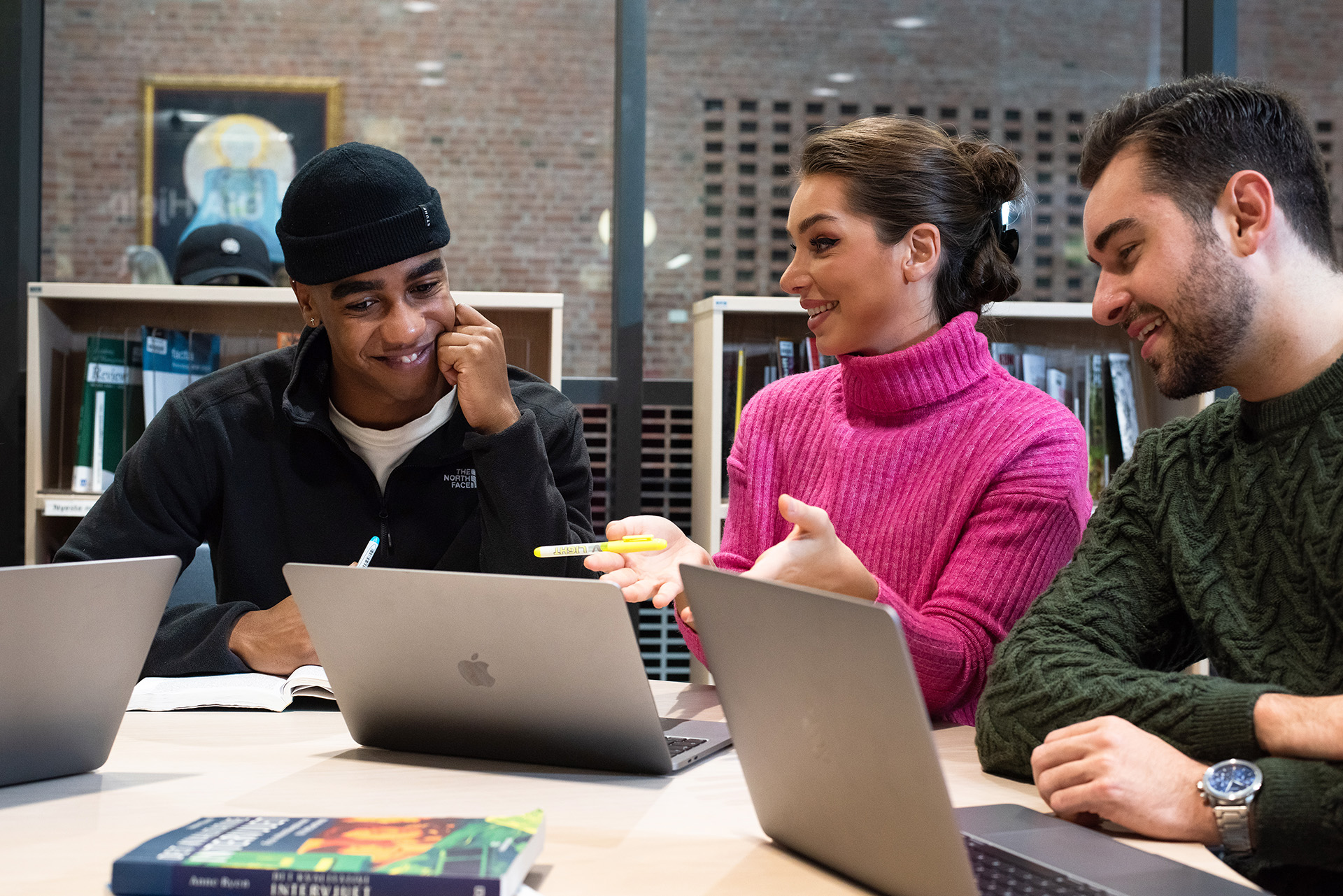 Tre smilende studenter sitter rundt et bord og jobber sammen med laptoper foran seg.