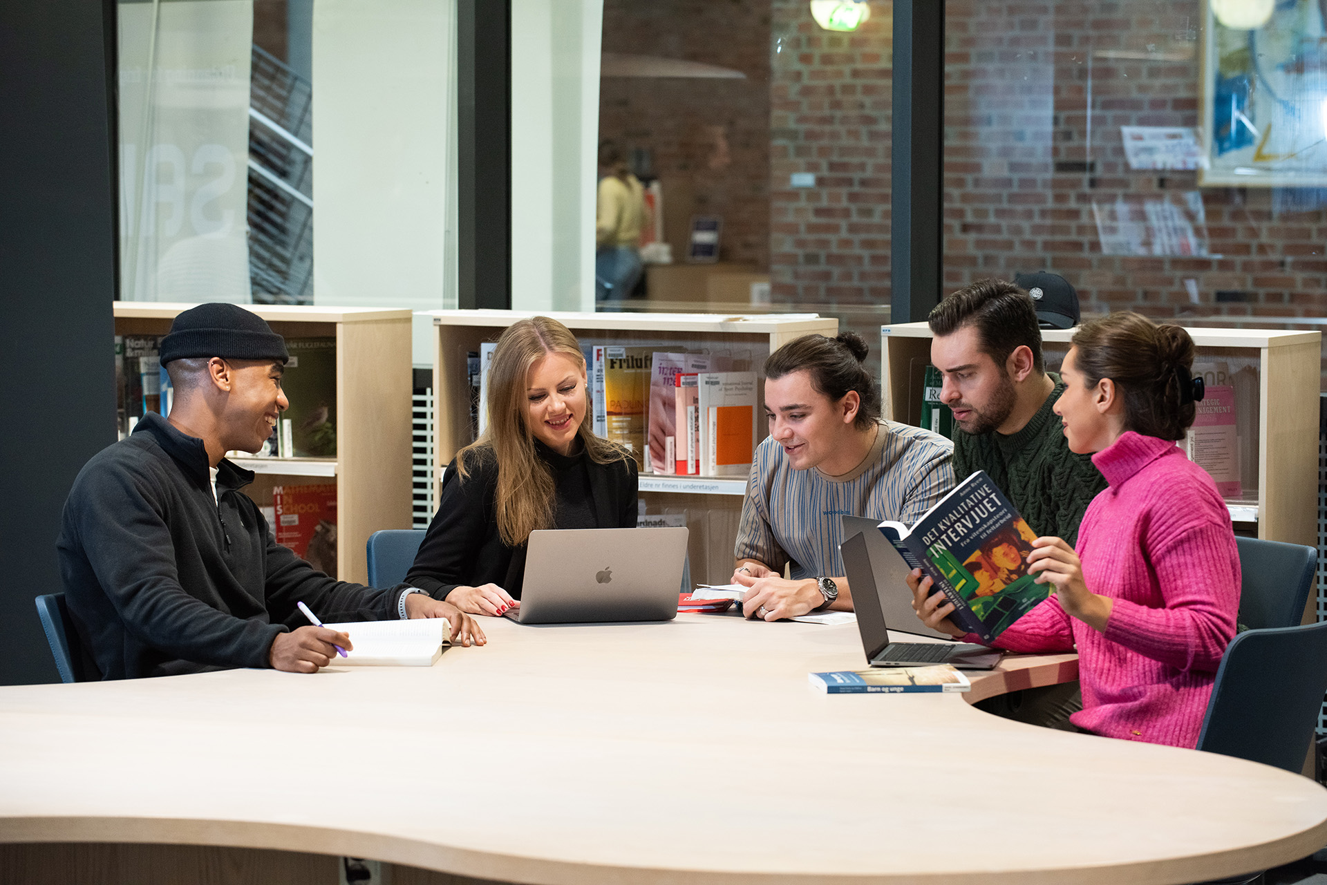 Fem smilende studenter sitter rundt et bord på et universitet og studerer.