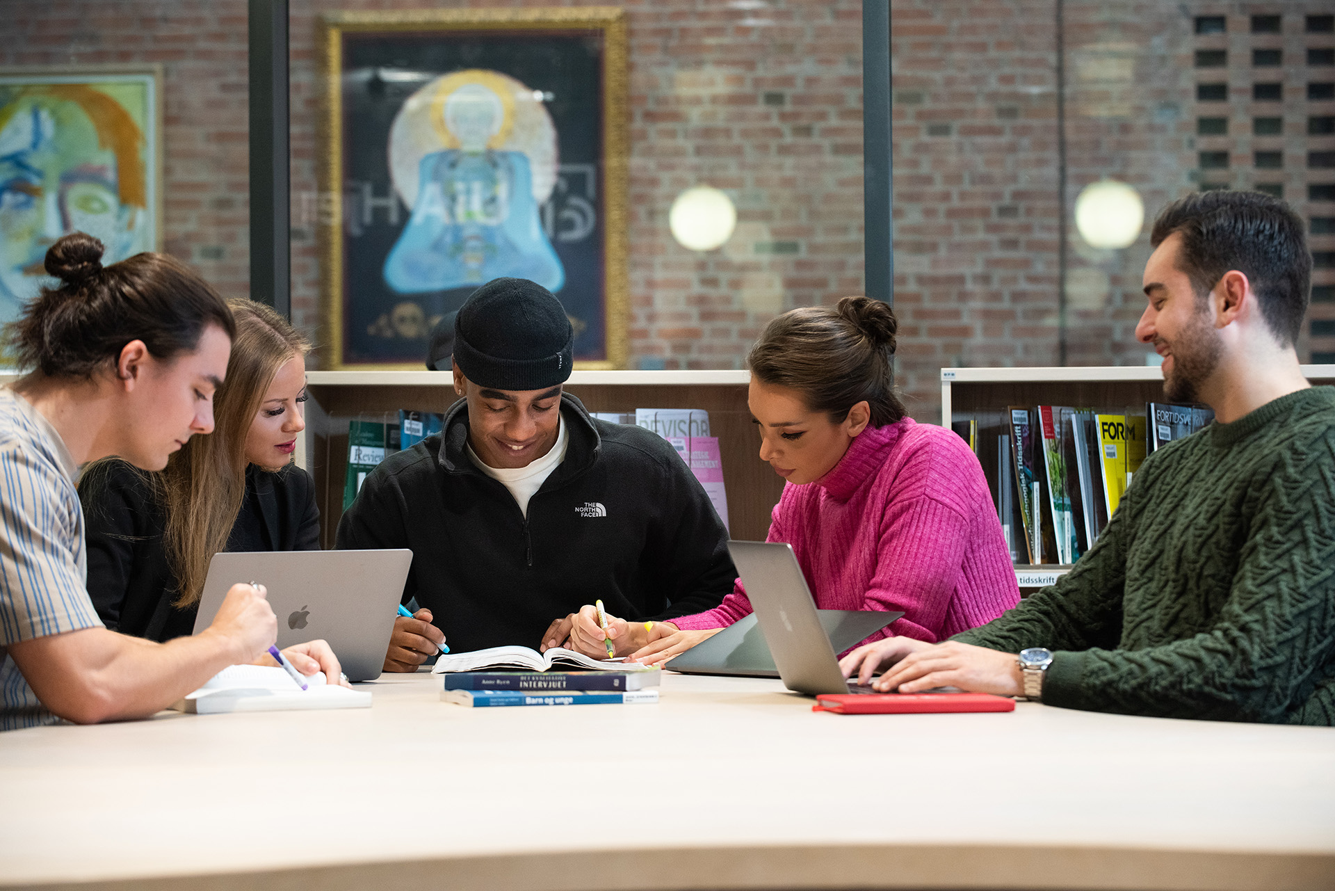 Fem smilende studenter sitter ved et bord, i et bibliotek, på et universitet og studerer.