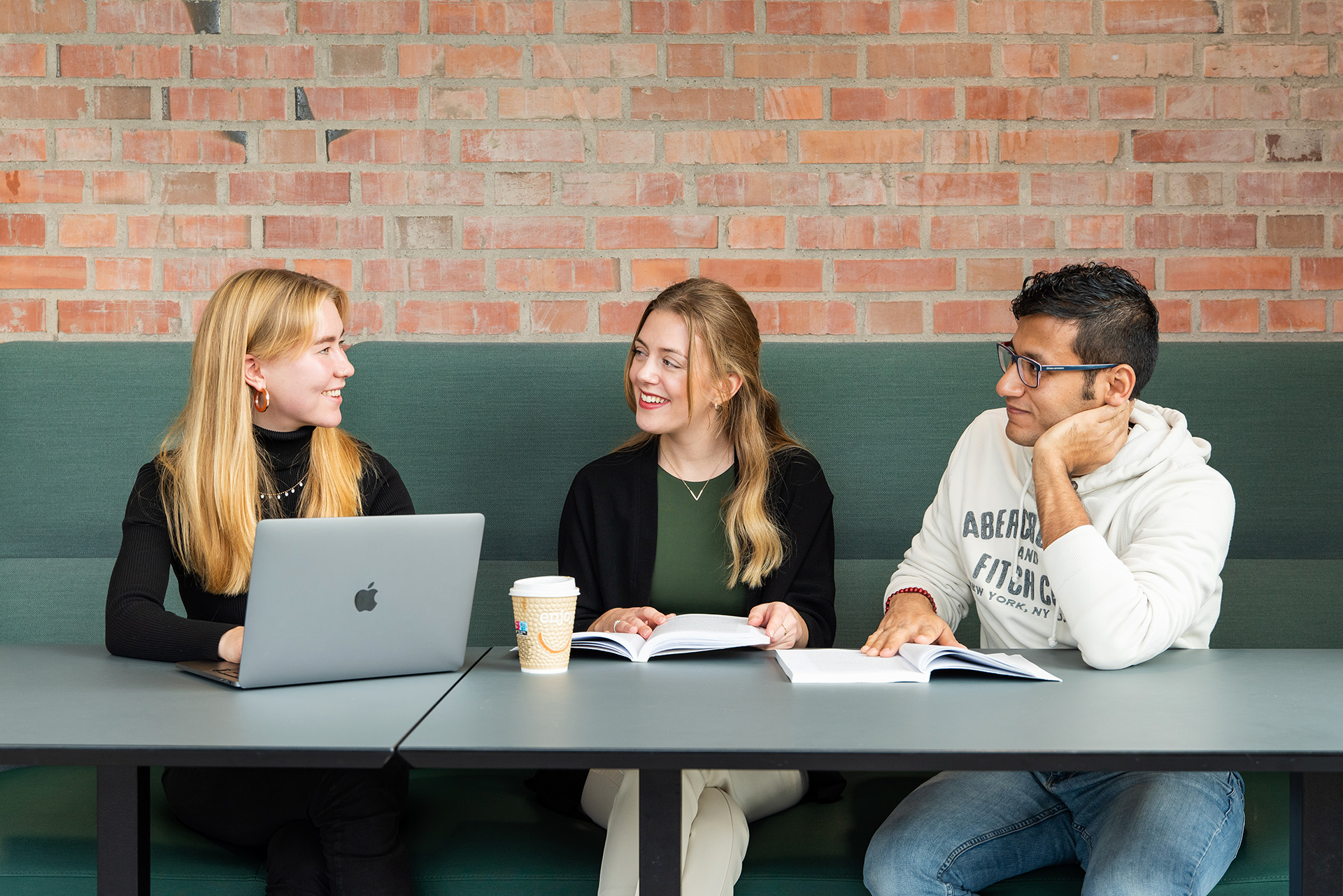 Tre smilende studenter sitter i en sofa ved et bord og snakker sammen over studiebøkene og en laptop.