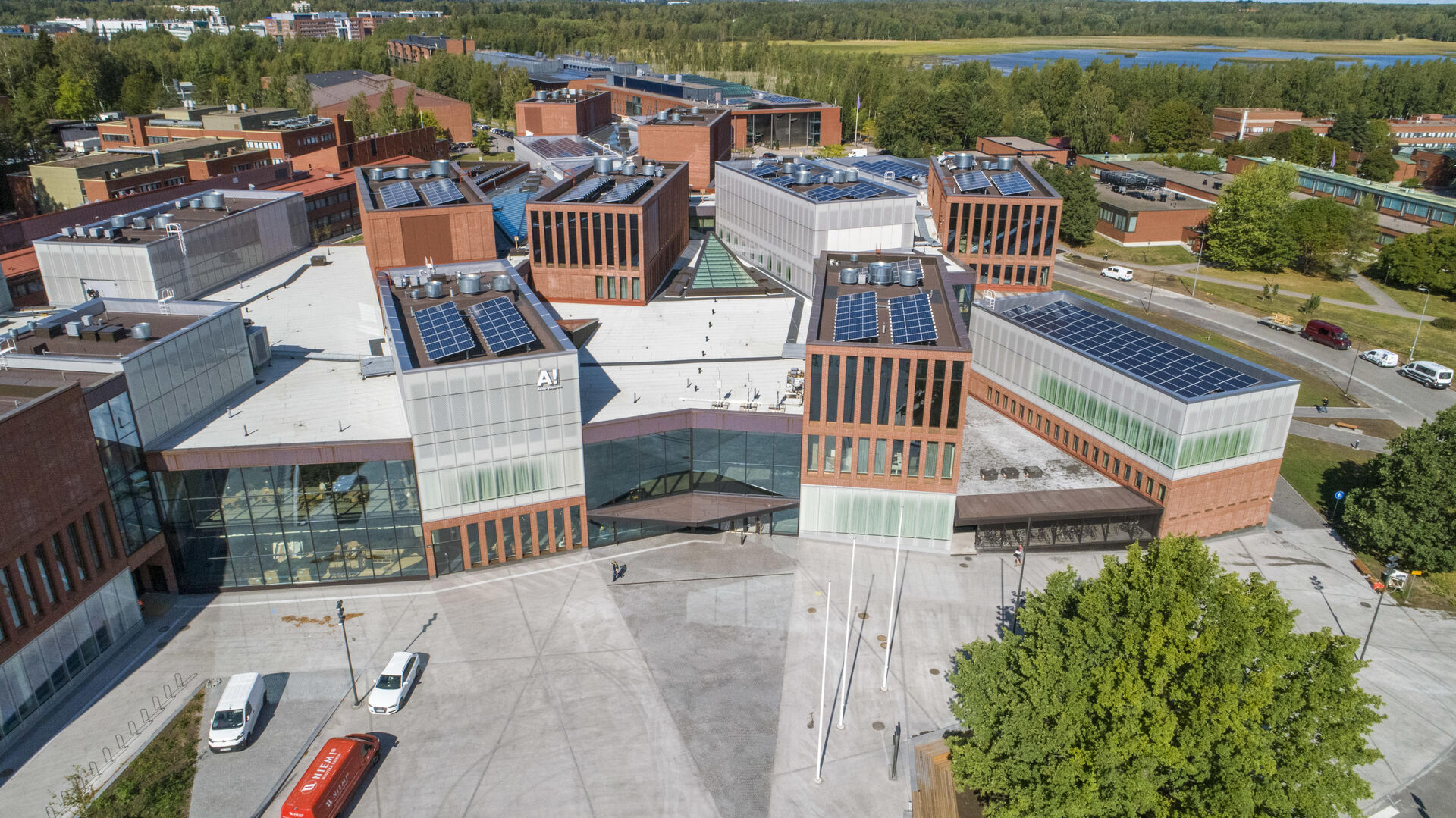 Dronefoto av Aalto University. Mange campusbygninger i rød murstein, betong og glass der man kan skimte trær og en innsjø i bakgrunnen.