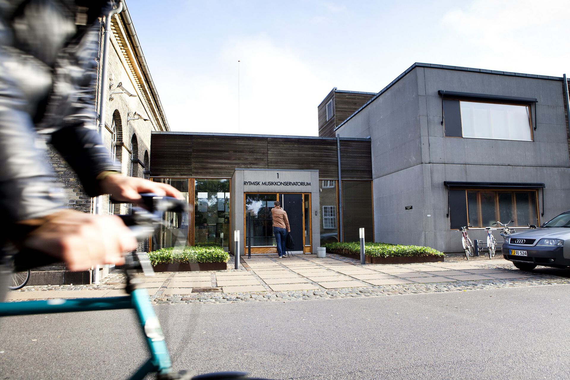 Hovedinngangen til skolen der det står Rytmisk musikkonservatorium over døra. En person suser forbi bygget på en blå sykkel.
