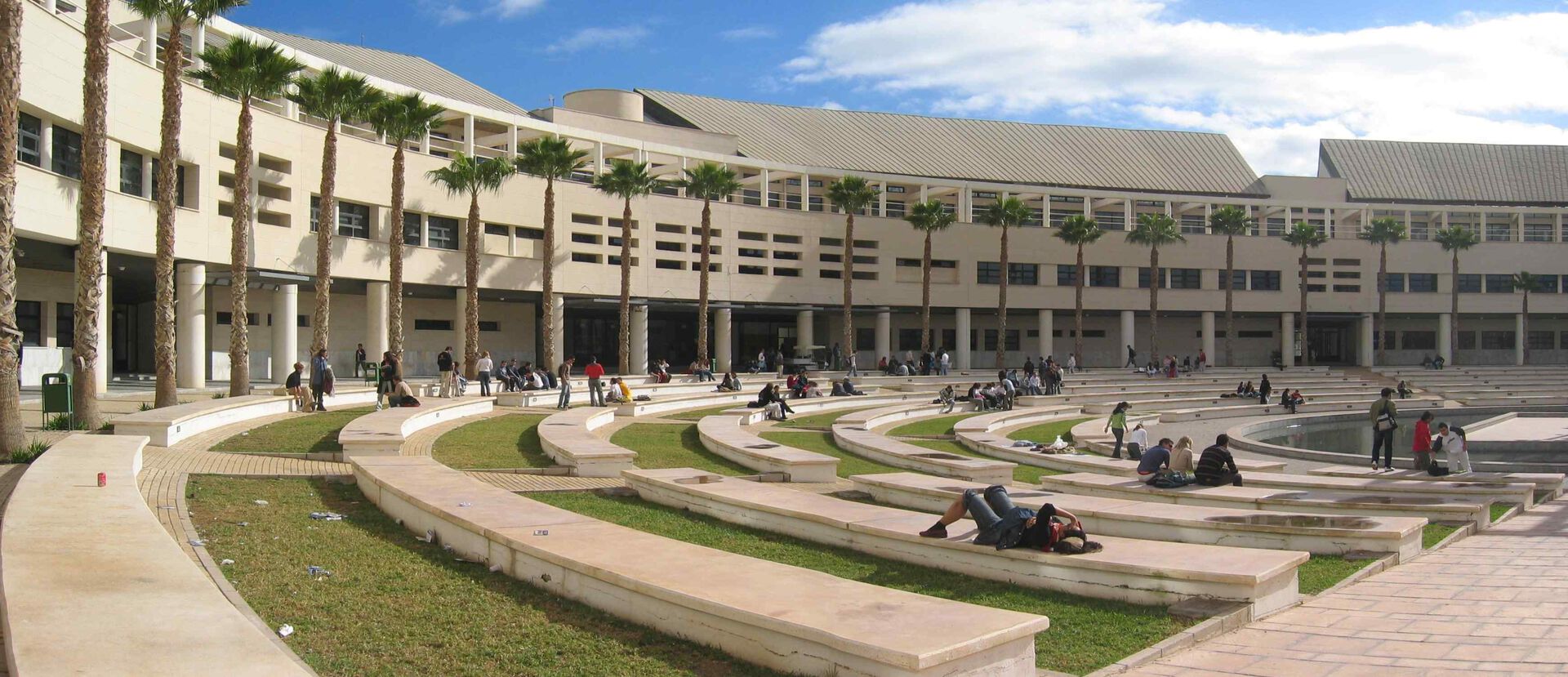 Campus Alicante med en beige, buet murbygning. Flere palmetrær og grønt arealer med benker i stein,